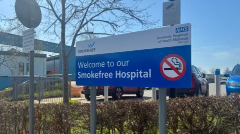 Local Democracy Reporting Service A mainly blue and white sign by a hedge includes the words "Welcome to our Smokefree Hospital" and a red, white and black no-smoking sign. Some cars are visible in the background.