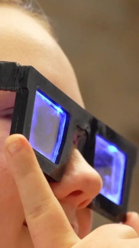 A close up image of a girls face, wearing thick rimmed black glasses which have blue glowing lenses