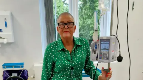 Tracie Mills Tracie having chemotherapy treatment, she wears a green shirt with white dots on it and has glasses on. Her head is shaved and she holds a pole in her left hand which has medical equipment on it