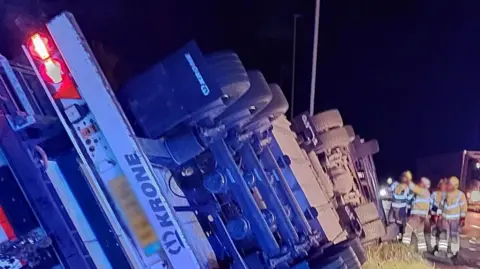 An overturned lorry at night, with emergency services nearby