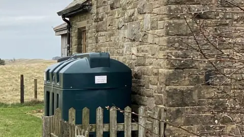 A green heating oil tank is positioned against a stone wall with a wooden fence in front of it.