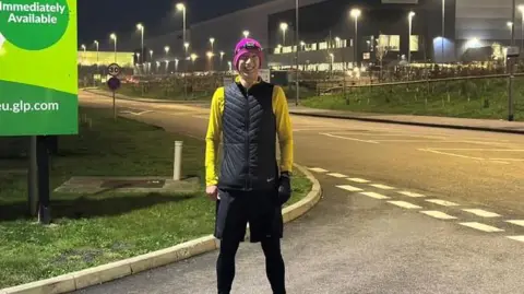 Ted Winterton Ted Winterton is stood in his running gear smiling. He is wearing a black overcoat with no arms, over a bright yellow fleece and is wearing a bright pink hat. The hat also has a headtorch round it. He also is wearing black running shorts over black thermal trousers. There is a large industrial building behind him and a road in front of that. 