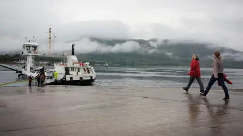 Two women walk down a slipway towards the MV Corran, a white and black ferry with some cars on its deck. It is a rainy day with low cloud on hills on the opposite shore of a loch.
