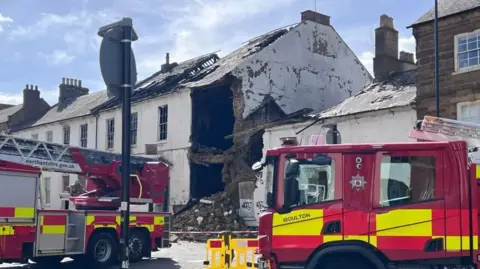 Kris Holland/BBC Two red and white fire engines at right angles to each other. They are parked in front of a two-storey white building that shows signs of fire. There is rubble in the street outside the houses. There are yellow barriers across the front of the fire engine on the right.