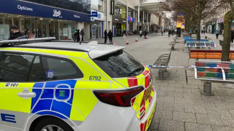 Police car and cordon around a number of benches in a pedestrian area of Southampton - Boots - the chemist shop on the left hand side.