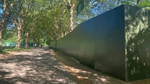 A large green fence running the length of a path in Sefton Park. People are walking in the distance.