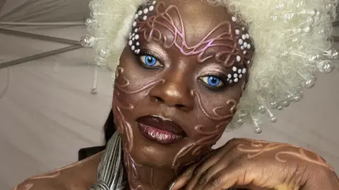 A person in bold avant‑garde makeup with bright blue eyes, a large white beaded wig, and metallic costume accessories, standing in a tent-like backstage setting