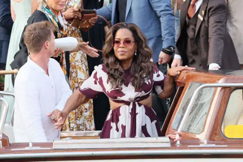 Getty Images Oprah Winfrey in Venice, Italy, for the wedding of Jeff Bezos and Lauren Sanchez. Oprah is wearing a dress featuring a burgundy and white print. Her hair has been styled in loose waves. 