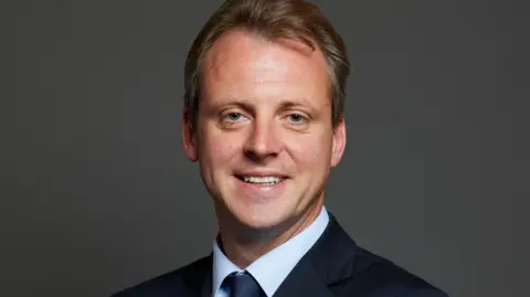 Joe Robertson poses for his official portrait as MP. He is looking at the camera, smiling and stands in front of a plain dark grey background. Mr Robertson has short blonde hair and is wearing a light blue collared shirt, with a navy blue tie and blazer.