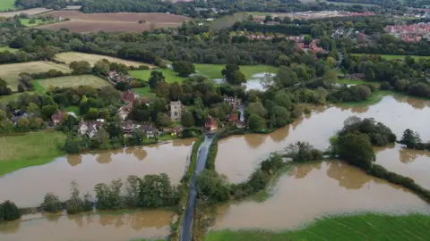 Dan Jessup An aerial view showing a flooded Sussex village with fields in the foreground that are completely underwater, a church and a few houses around it are also seen while in the background there are more homes, tree and other fields, some with flooding.