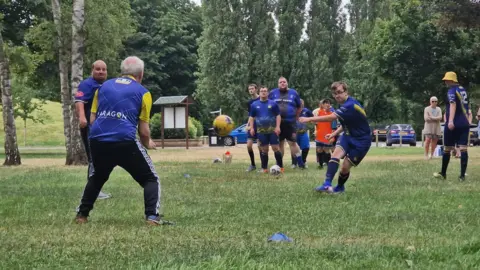 Naj Modak Several football players in blue kits take part in a training drill on a grass field in a park. Taylor kicks a yellow football while teammates and spectators watch from nearby.