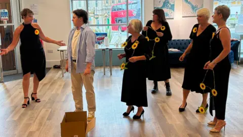 Teylu Theatre Company Four women wearing black with sunflower necklaces and broaches are performing a scene from Calendar girls 