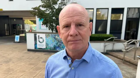 Richard looks at the camera with a serious expression. He wears a light blue shirt and is front of a building with the Bristol Water logo on it. 