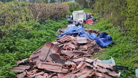 Many broken roof tiles are piled on the floor of a grassy bridleway. Further down is a blue tarp covering more waste, and also a white sofa.