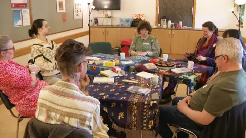 Seven people sit around a table. Various books and biscuits are laid out in front of the group, who are talking and laughing with each other