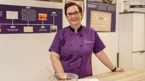 Geri Martin Geri Martin - a woman wearing a purple chef's coat with 'The Chocolate Manor' embroidered in white. She stands at a wooden kitchen island with a bowl of chocolate in front of her.