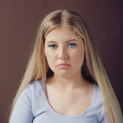 Museum Wales Head and shoulders shot of a girl with long blonde hair. She wears a scoop neck light blue cotton top and has striking eye make up on, with electric blue eye liner underneath and light purple eyeshadow on her lids. She looks at the camera without smiling.