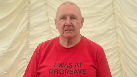 Robert Young, a man in his 80s with shaved hair, is wearing a bright red shirt which reads in black lettering: "I was at Orgreave."