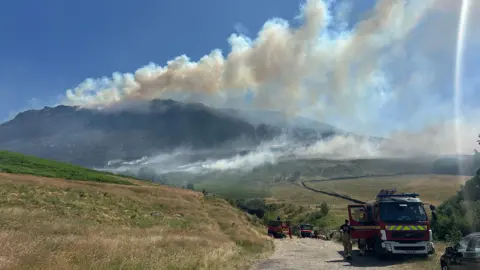 Three fire engines are parked up on a dirt track on the moors, with plumes of smoke seen billowing from the moors in the distance.
