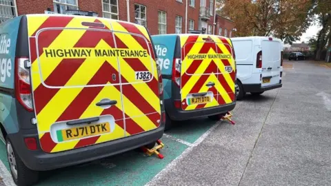 Local Democracy Reporting Service Two vans which say 'Highway Maintenance' with clamps on the rear back wheel. They are in a car park.