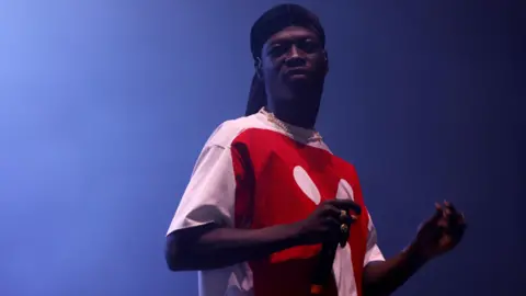 A man with a black head covering, gold chain and red and white shirt, holds a microphone while performing on stage.