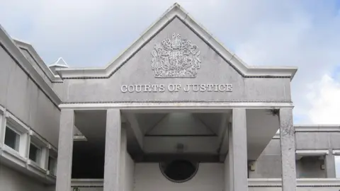 The front of Truro Crown Court. It is grey stone building with COURTS OF JUSTICE written below an elaborate heraldic shield. There are four pillars and the roof is pitched to a point.