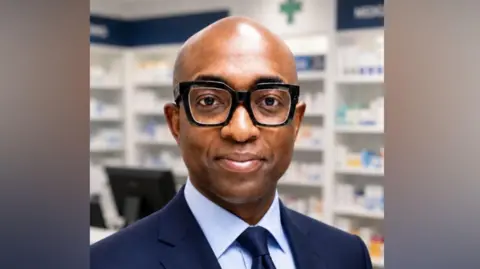 Ade Williams Ade in a mid-shot standing in a pharmacy with shelves of medicines behind him. He is a black man and is bald with black glasses. He is wearing a smart navy suit jacket and tie with a pale blue shirt .
