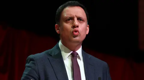 Anas Sarwar speaking at an STUC event in April 2025. He is wearing a dark blue blazer, a white shirt and a purple tie and is standing on a stage with a red curtain backdrop.