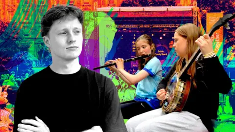 Jamie McColgan in a black tshirt with his arms folded, there are some people playing instruments to his right. 