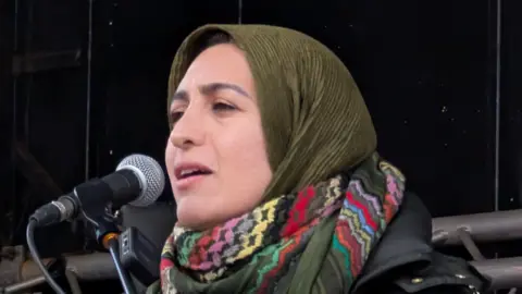 Azadeh Sobout is wearing a green hihab and a multi-coloured scarf as she speaks into a mic.