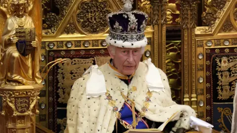 King Charles sitting on a golden throne with a crown on. 