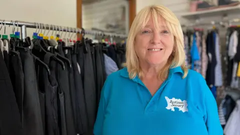 Lesley stands in a clothing store wearing a blue polo shirt with the text 'Autism Jersey.' Behind them are racks of pants and shirts on hangers, shelves with folded clothes, and decorative items.