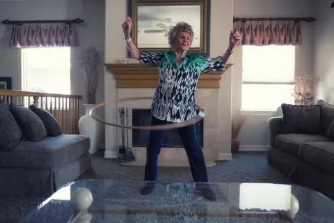 Chris Reese and Amy Hill Joan in a pair of dark dark jeans in the Aztest shirt while spinning with Holly collar around her waist in a hall with brown sofa, gray carpet and a mirror over a cloak