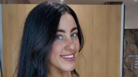 Tia, a young woman, is smiling with her teeth at the camera. She has bright blue eyes and black hair in a side parting and looks at the camera from the side. 