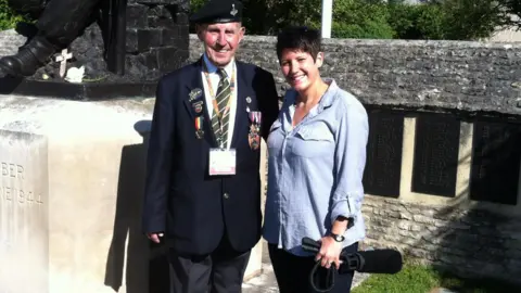A man wearing military medals stands next to a woman wearing a grey shirt and holding a microphone. They are standing in front of a large war memorial which shows a solider with a helmet and gun. In the background are lists of names. The memorial says "Remember the 6th June, 1944".