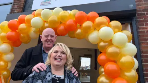 Claire Hamilton/BBC Steve Herron poses with his hands on Sue Herron's shoulders standing in front of an archway made of orange, golden and yellow balloons 