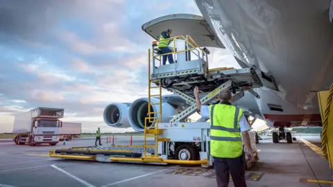 Two ground crew members in hi-vis jackets load an aircraft on the runway. 