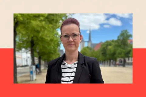 A white woman with purple hair swept back off her face and glasses. She is wearing a dark blazer and black-and-white striped top, and is looking at the camera
