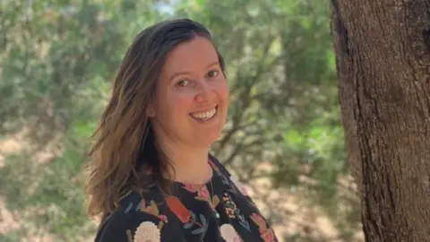 Dr Emma Lawrance standing beside a large tree wearing a dark floral-patterned top. She has long brown hair and is smiling. The scene is outdoors in a wooded area.