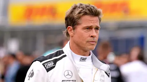 Brad Pitt with light brown hair and blonde highlights and wearing a white Mercedes F1 racing suit.