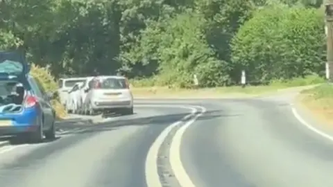 Liz Gillham Cars parked on a road with a blind bend and double white lines down the middle. There are trees and hedgerows at the side of the road.