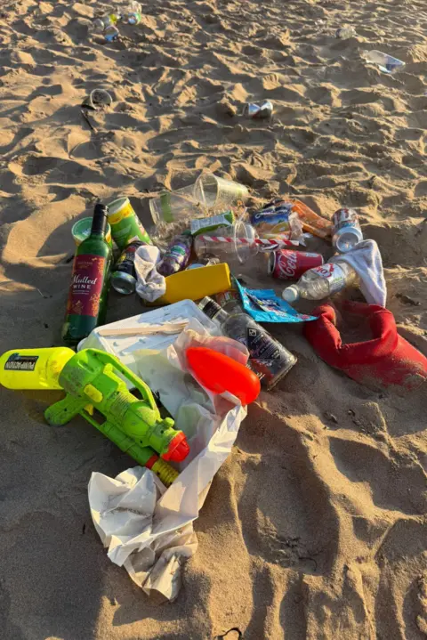 Lisa Wallace Bottles, cans, plastic cups and takeaway wrappers covered in sand. A neon green water gun is beside the rubbish covered in sand. 