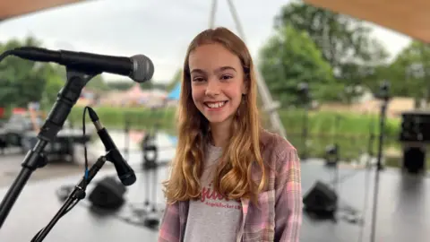 Anna, an 11-year-old girl, standing on a stage with a microphone, smiling at the camera. There are trees and a lake behind her.