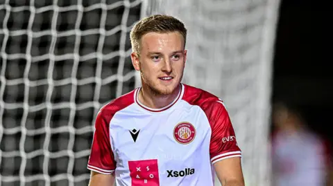 Harvey White smiles looking ahead with sweat on his brow, wearing home Stevenage kit of white and red