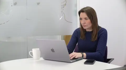 JAMIE NIBLOCK/BBC Nikki Fox sits at a desk wearing a deep purple woollen dress. There is a frosted glass wall to her right behind her and a white cup to her right on a white desk. Her silver laptop sits in front of her, while a black phone is on the large desk to her left. She is looking at a screen and concentrating on the conversation.