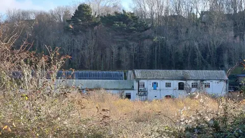 A white derelict mill building on a piece of scrubland. A wooded hill rises up behind it and houses can be seen through the trees. 