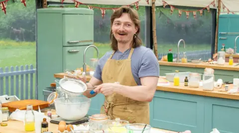 Channel 4 Iain Ross has shoulder length auburn hair and a moustache. He is wearing dangly earrings and necklace and a blue jumper and camel coloured apron. He is in the Bake Off tent standing at a table sifting icing sugar into a bowl.