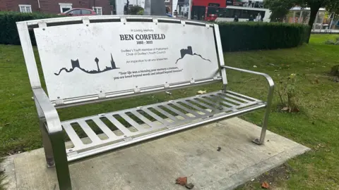 A silver memorial bench in a park, which says "Ben Corfield 2003-2022" with inscriptions underneath