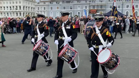 Three men dressed in military regalia holding drums on Plymouth Hoe.
