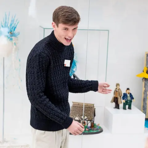 Lego House Kit Nugent, who has brown hair and is wearing a navy jumper and beige tousers, points to his models in a glass case during the opening of the exhibition at the Lego House. 